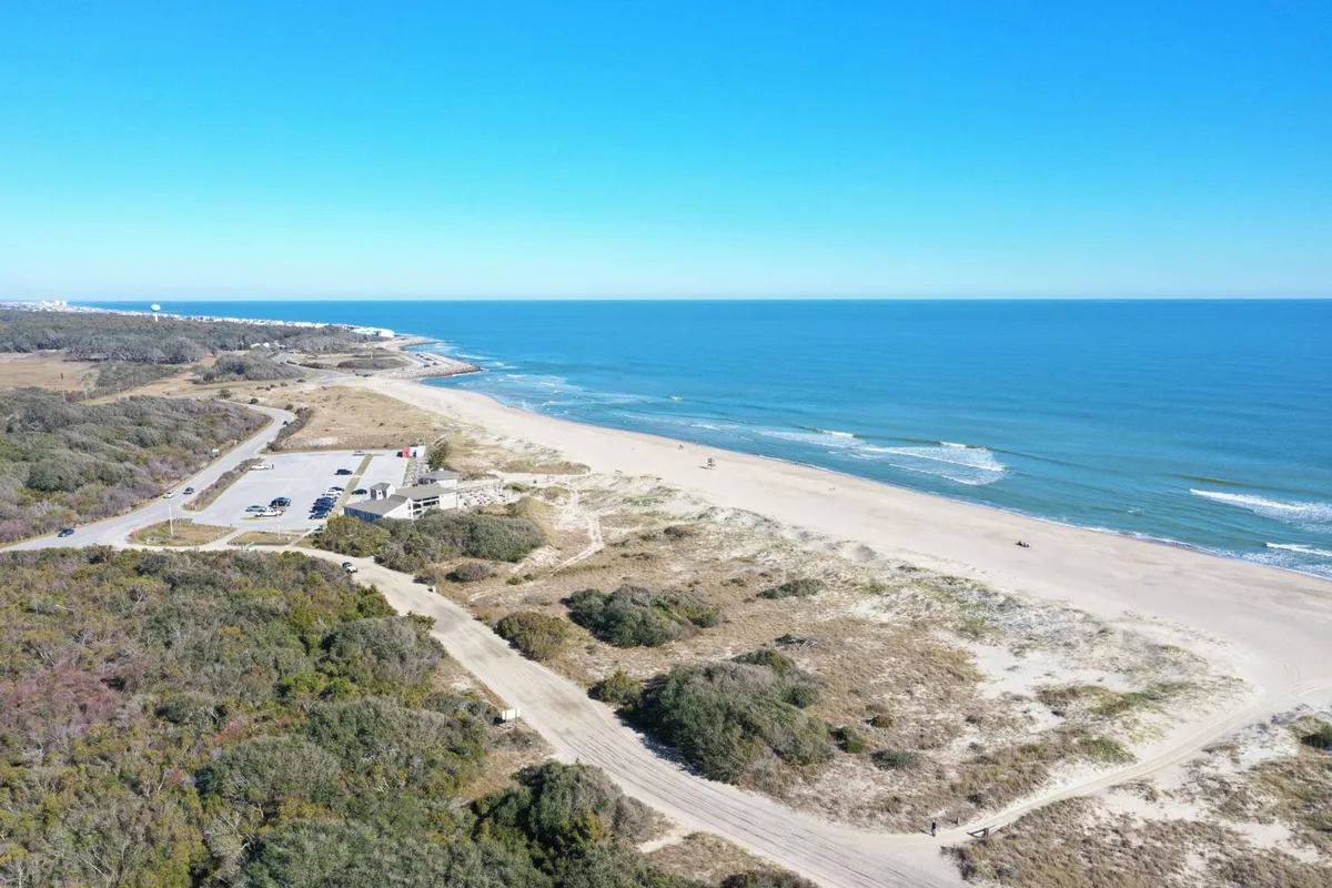 A tranquil scene showing the ocean, sandy beach, parking area, and grassy dunes. The area appears serene and ideal for sunrise or sunset walks and enjoying the natural beauty of the coast.