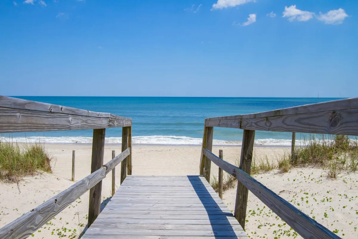 This image captures a wooden walkway leading to the beach, with the ocean and expansive sky in the background. The scene evokes a sense of calm and anticipation, perfect for early morning strolls or group gatherings on the sand. The relaxed atmosphere is enhanced by natural light and the openness of the coastline.