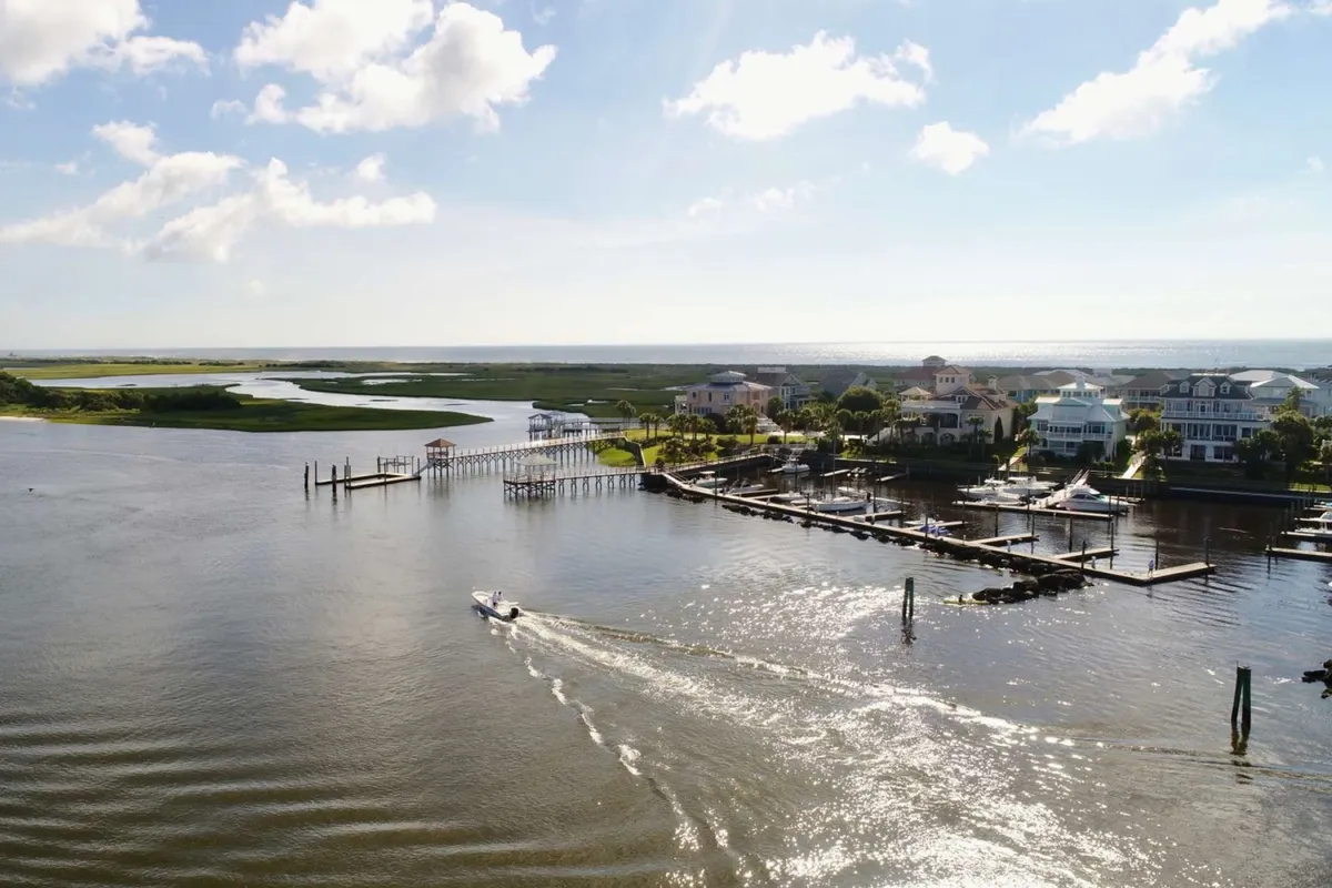A scenic view of a river with boats docked along wooden piers, houses on the shoreline, and a wide sky overhead. The tranquil water invites thoughts of boating excursions, waterfront relaxation, and exploration of the surrounding area.