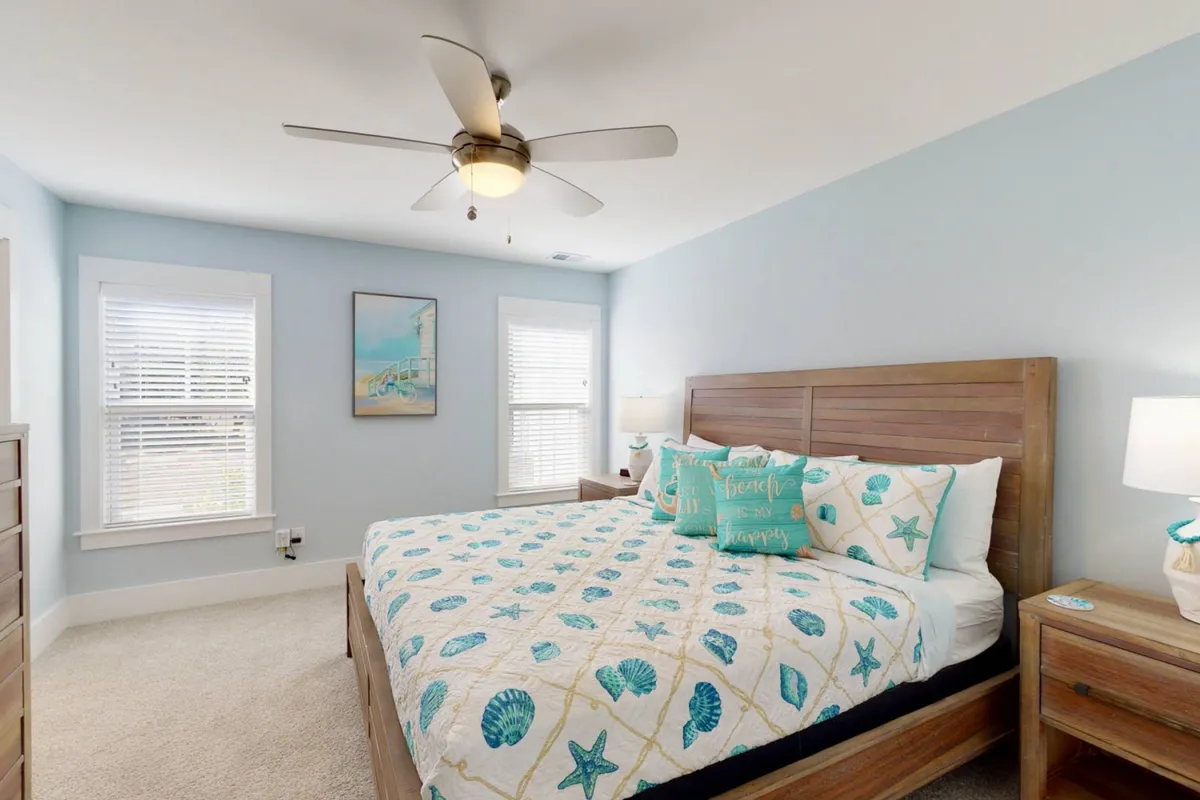 A tranquil king bedroom featuring a neatly made bed with decorative pillows, framed artwork above the headboard, and bedside tables with lamps that cast a soft, calming glow. Natural light spills in, creating a soothing and restful atmosphere.