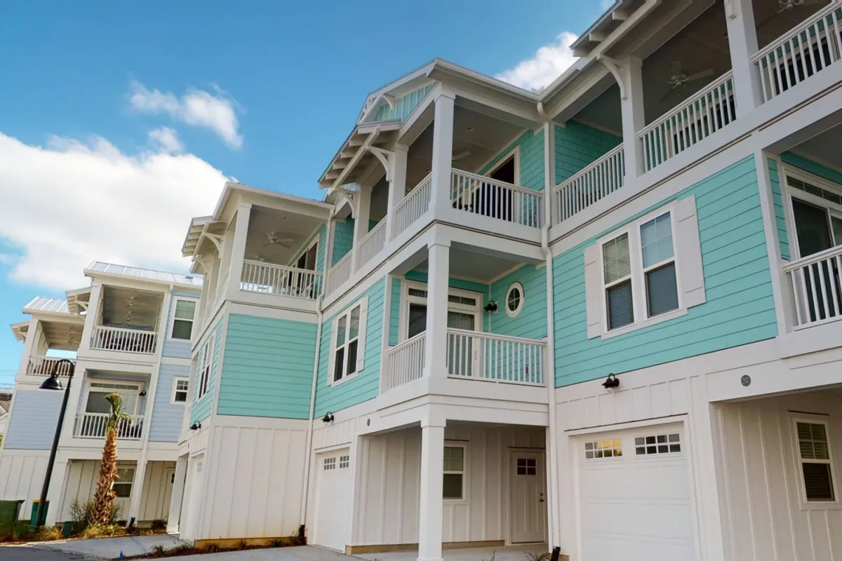 A cheerful exterior view of Barefoot Cove's pastel-colored townhomes, complete with balconies, architectural details, and visible gathering spaces. The scene captures a sunny, inviting neighborhood vibe with opportunities for socializing and outdoor enjoyment.