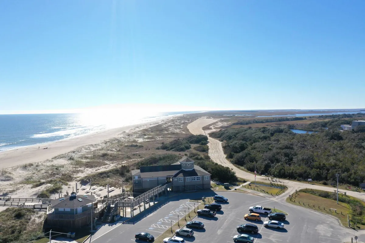 A sweeping exterior view of the shoreline at Kure Beach, showcasing the beachfront, a parking area, a coastal path, and distant dunes. The sky is clear, and the ocean stretches out invitingly, emphasizing a relaxed beach lifestyle.