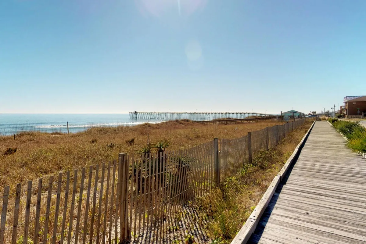 The view showcases a scenic pier extending into the ocean with a sandy beach, boardwalk, and dune grass. The composition is lively yet serene, suggesting activities like building sandcastles, fishing off the pier, and family walks along the shore. The pier depicted could easily represent the iconic Kure Beach Pier mentioned in the text.