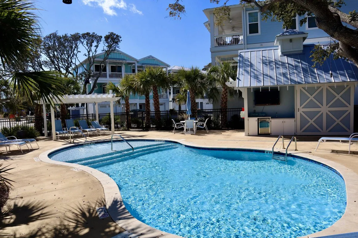 The image showcases the inviting outdoor amenities of Barefoot Cove: a sparkling swimming pool surrounded by comfortable lounge chairs, swaying palm trees, and a stylish outdoor kitchen area. The scene is set for relaxation, social gatherings, and enjoying the coastal Carolina sunshine, capturing the essence of resort-style living.